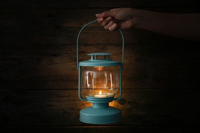 Woman holding lantern with burning candle against wooden wall in darkness, closeup