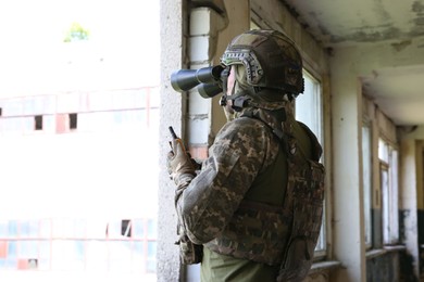 Photo of Military mission. Soldier in uniform with binoculars inside abandoned building, back view. Space for text