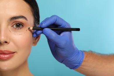 Doctor with pencil preparing patient for cosmetic surgery operation on light blue background, closeup