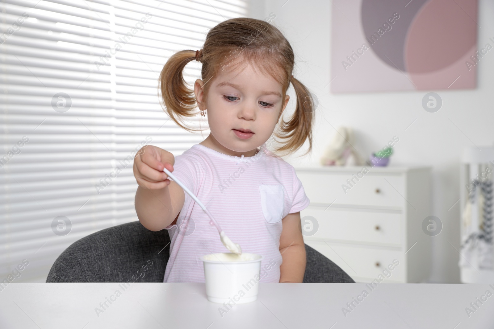 Photo of Cute little child eating tasty yogurt from plastic cup with spoon at white table indoors