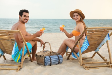 Happy young couple with cocktails sitting on deck chairs at sea beach