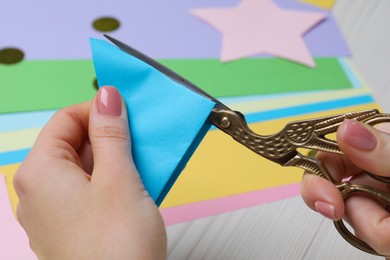 Woman cutting light blue paper with scissors at white wooden table, closeup