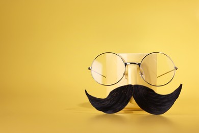 Man's face made of artificial mustache, glasses and cup on yellow background. Space for text