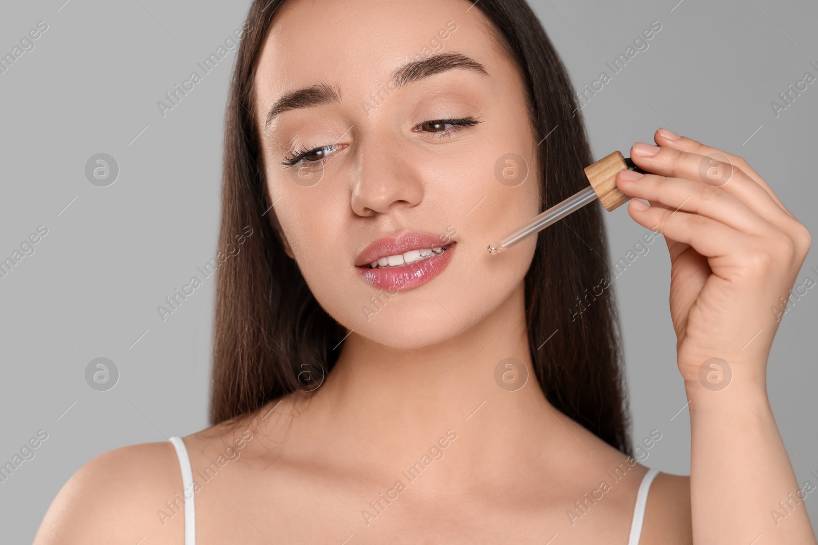 Photo of Beautiful young woman applying essential oil onto face on grey background