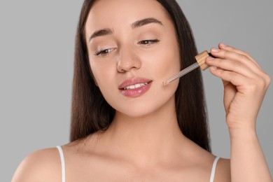 Beautiful young woman applying essential oil onto face on grey background