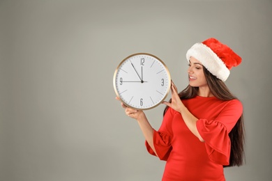 Young beautiful woman in Santa hat holding big clock on grey background. Christmas celebration