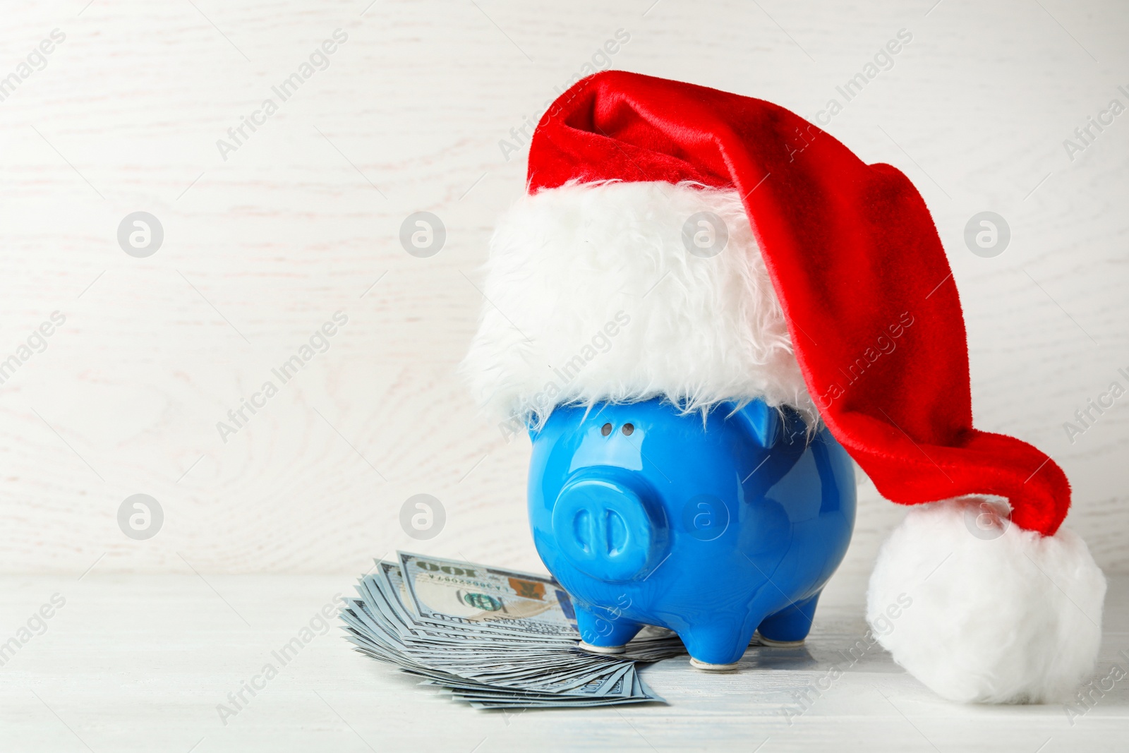 Photo of Piggy bank with Santa hat and dollar banknotes on white wooden table. Space for text