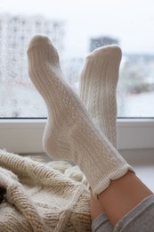 Photo of Woman in knitted socks relaxing near window at home, closeup