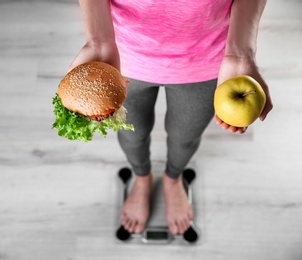 Photo of Woman holding tasty sandwich and apple while measuring her weight on floor scales, top view. Weight loss motivation