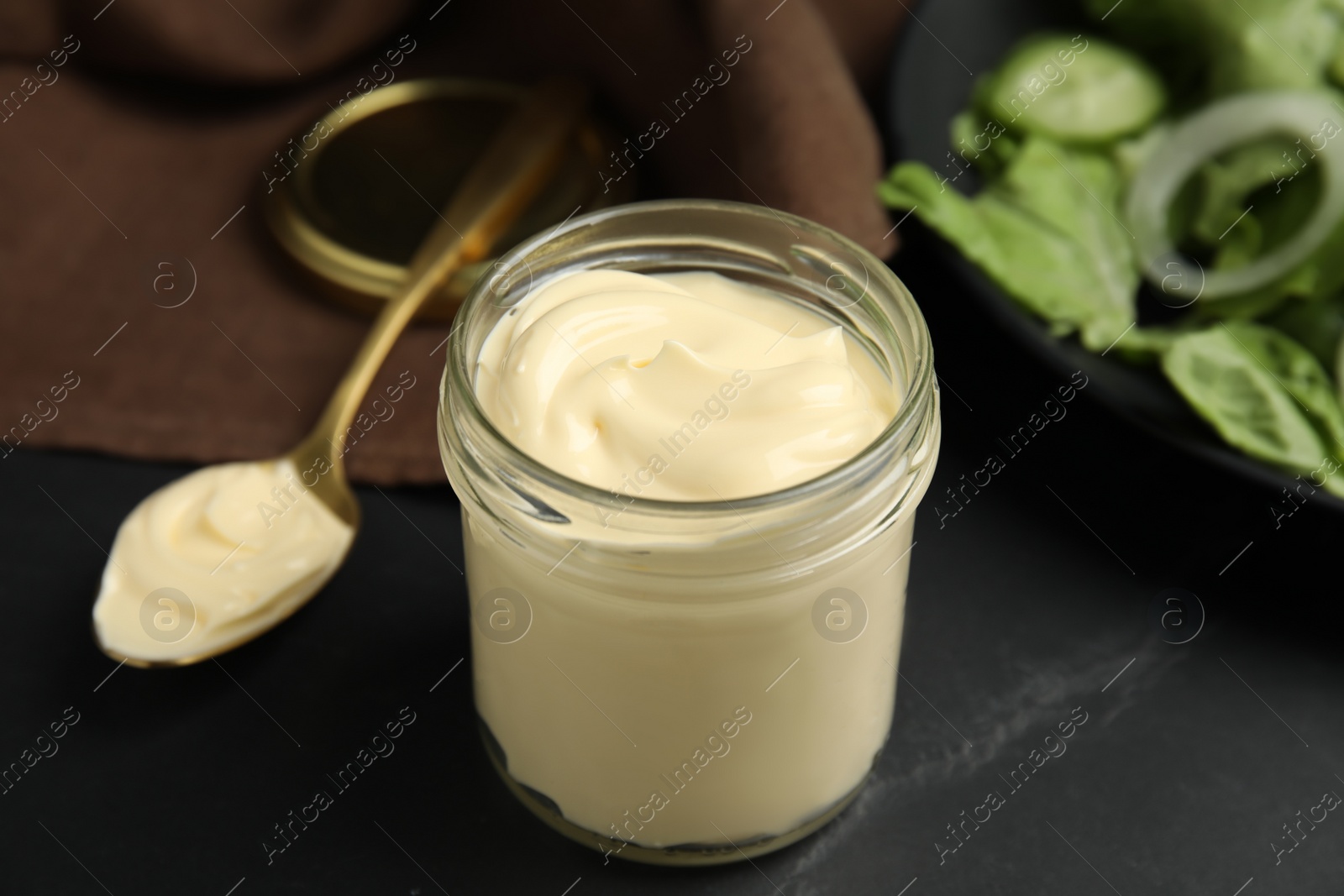 Photo of Jar and spoon with delicious mayonnaise near fresh salad on black table