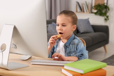 Little girl eating biscuit while using computer at table in room. Internet addiction
