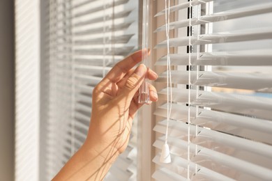 Photo of Woman opening horizontal blinds on window indoors, closeup