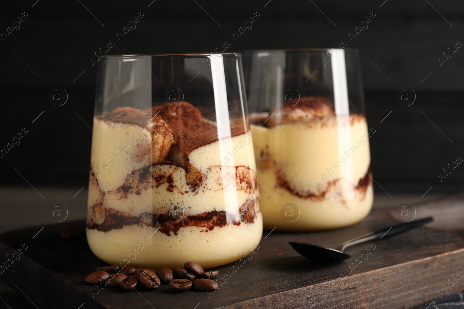 Photo of Delicious tiramisu in glasses, spoon and coffee beans on wooden board, closeup