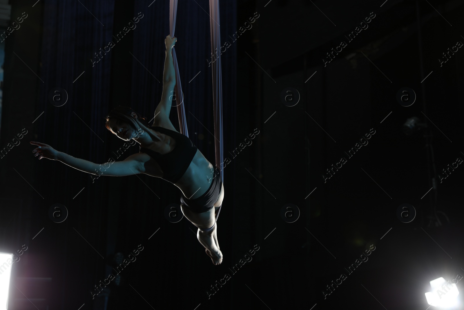 Photo of Young woman performing acrobatic element on aerial silk against dark background. Space for text