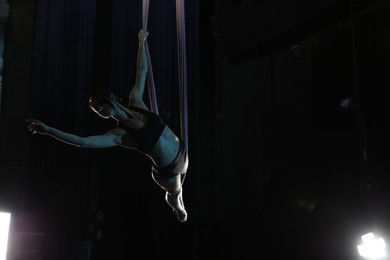 Young woman performing acrobatic element on aerial silk against dark background. Space for text