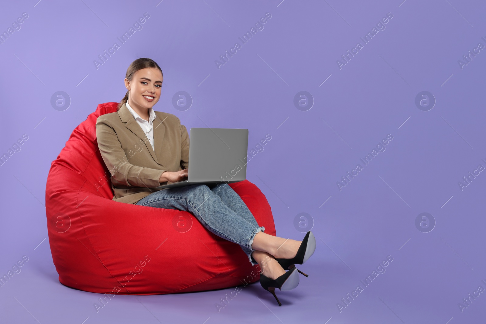 Photo of Happy woman with laptop sitting on beanbag chair against violet background. Space for text