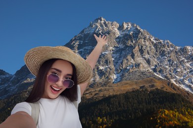 Smiling young woman in sunglasses and straw hat taking selfie in mountains