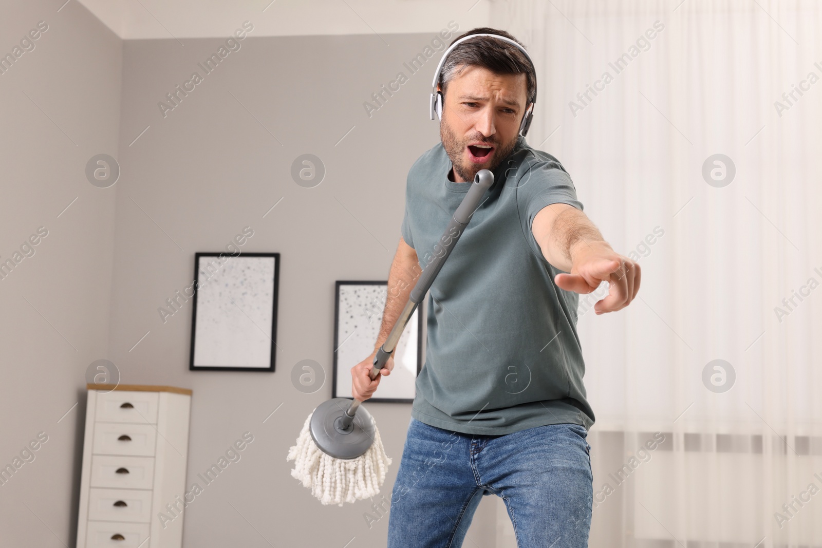 Photo of Enjoying cleaning. Happy man in headphones with mop singing while tidying up at home