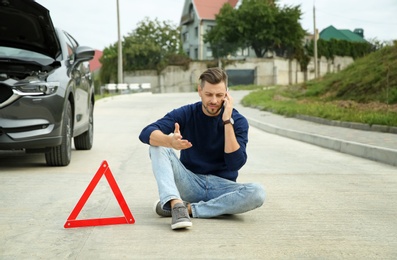 Photo of Man talking on phone near warning triangle and broken car on road