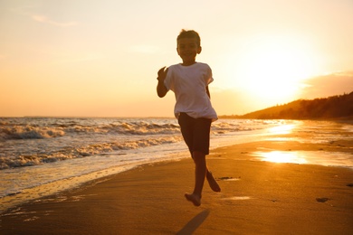 Cute little boy running on sandy beach near sea at sunset