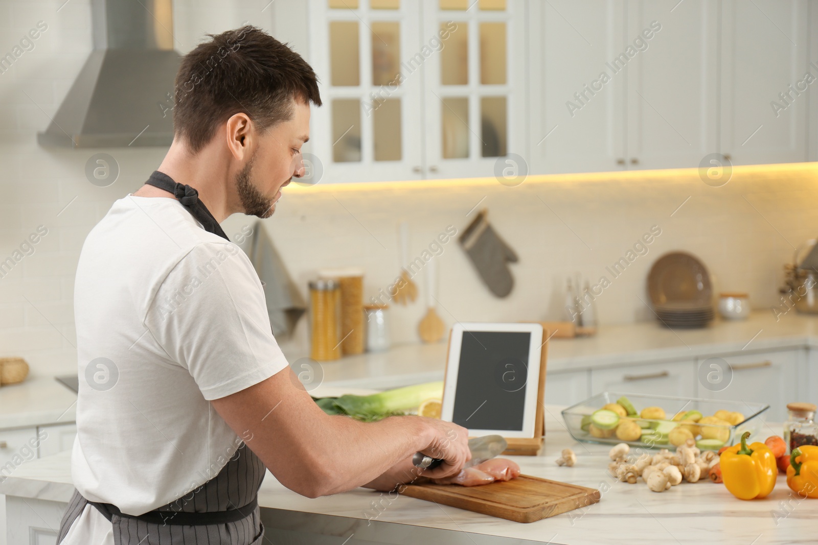 Photo of Man cutting chicken fillet while watching online cooking course via tablet in kitchen