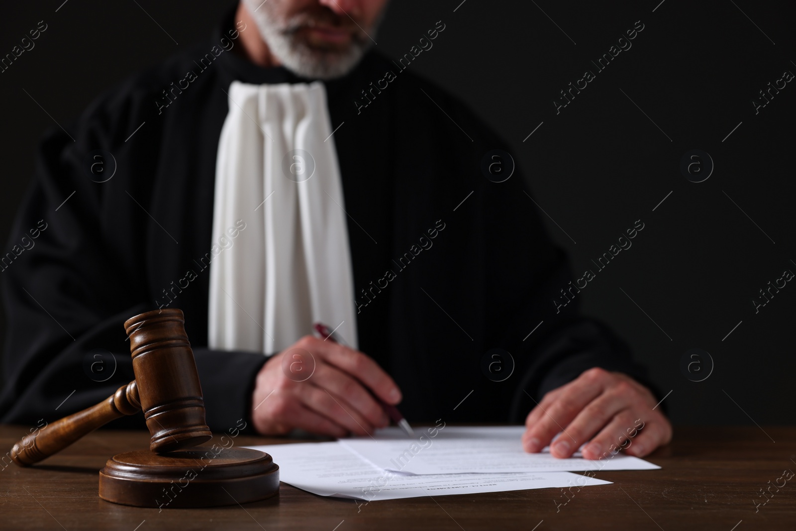 Photo of Judge with gavel writing in papers at wooden table against black background, closeup