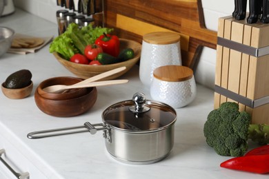 Metal saucepan, other cooking utensils and fresh vegetables on countertop in kitchen