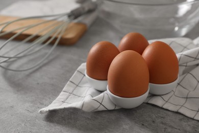 Photo of Chicken eggs, whisk, glass bowl, napkin and wooden board on grey table, space for text