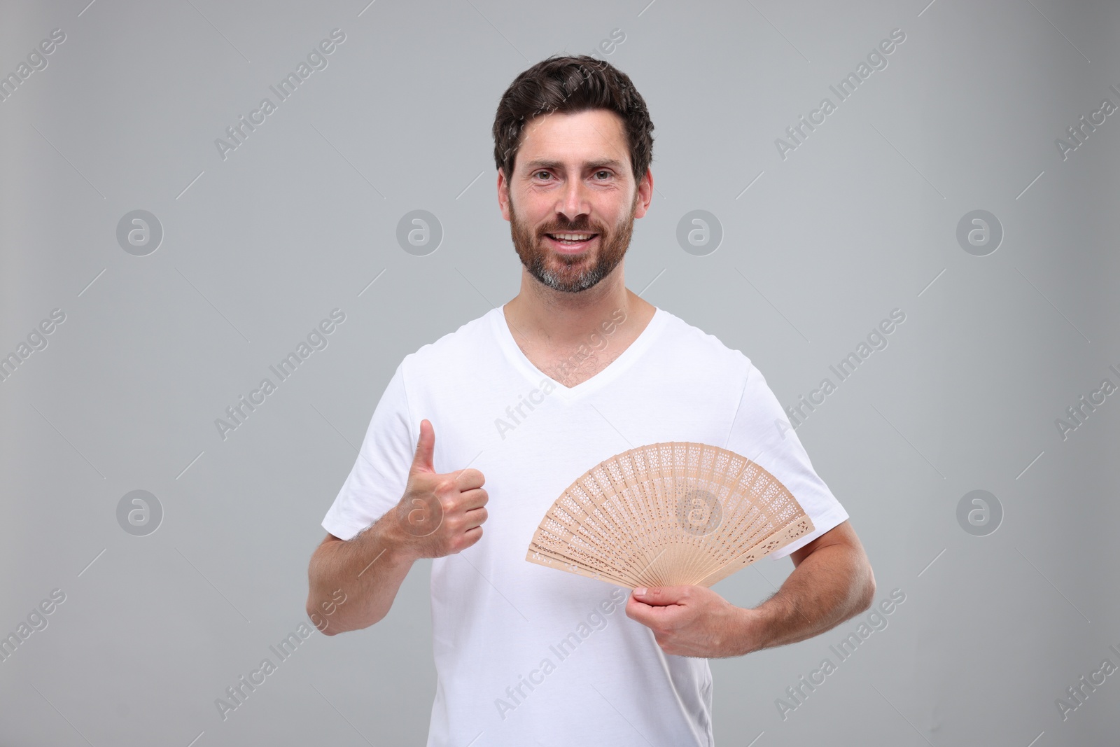 Photo of Happy man holding hand fan and showing thumb up on light grey background