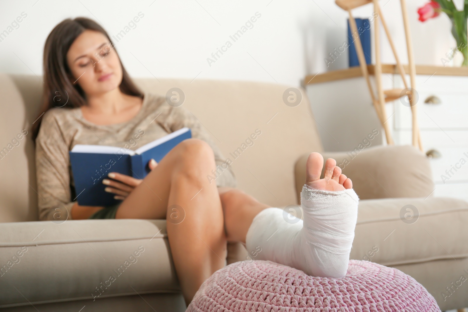 Photo of Young woman with broken leg in cast reading book while sitting on sofa at home