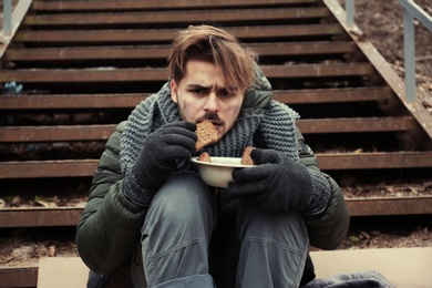 Photo of Poor young man with bread sitting on stairs outdoors