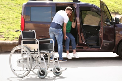 Photo of Young man helping woman to get out from van,  focus on wheelchair outdoors