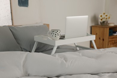 Photo of White tray table with laptop and cup of drink on bed indoors