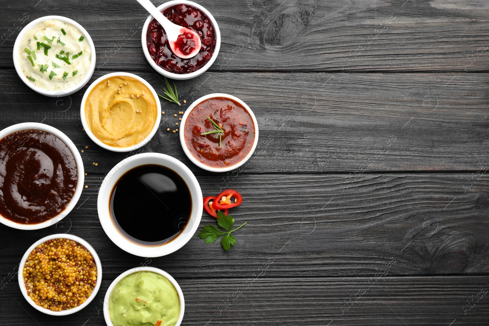 Photo of Different tasty sauces in bowls, parsley, chili pepper and rosemary on black wooden table, flat lay. Space for text