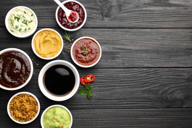 Photo of Different tasty sauces in bowls, parsley, chili pepper and rosemary on black wooden table, flat lay. Space for text