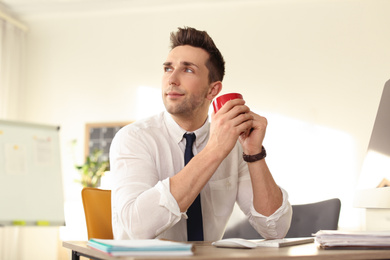 Young businessman with cup of drink relaxing at table in office during break