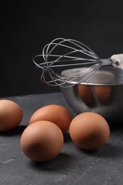 Photo of Metal whisk, raw eggs and bowl on dark grey table, closeup
