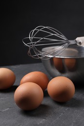 Metal whisk, raw eggs and bowl on dark grey table, closeup