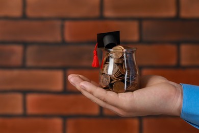 Man holding glass jar of coins and graduation cap against brick background, closeup with space for text. Scholarship concept