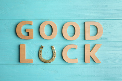 Phrase GOOD LUCK made of wooden letters and horseshoe on light blue table, flat lay