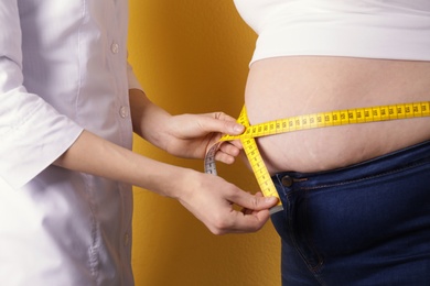 Doctor measuring waist of overweight woman on color background, closeup