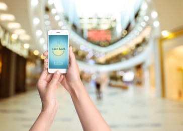 Image of Woman holding smartphone with word Cashback and shopping cart icon indoors, closeup. Space for text