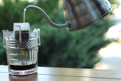 Photo of Pouring hot water into glass with drip coffee bag from kettle on wooden table, closeup