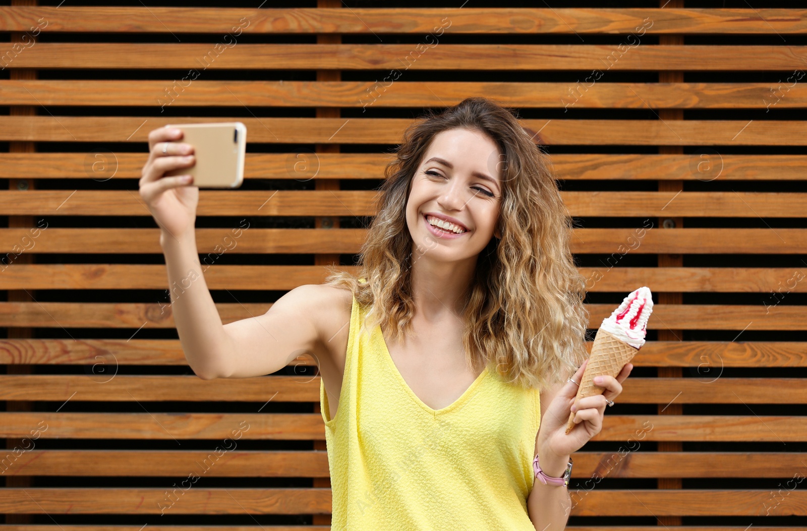Photo of Happy young woman with delicious ice cream in waffle cone taking selfie near wooden wall
