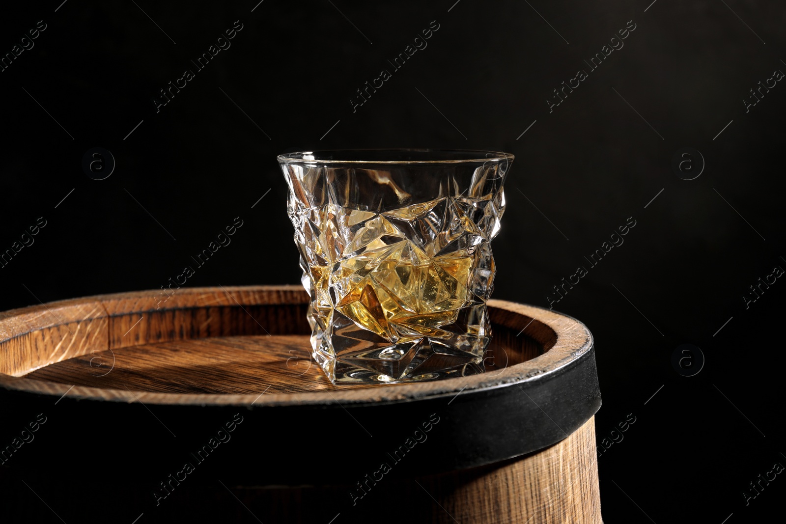 Photo of Glass of whiskey on wooden barrel against black background, closeup