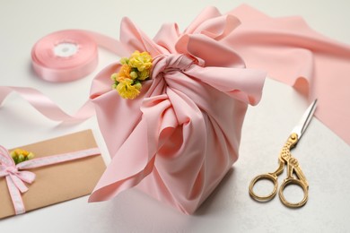 Photo of Furoshiki technique. Gift packed in pink fabric, card, flowers and scissors on white table, closeup
