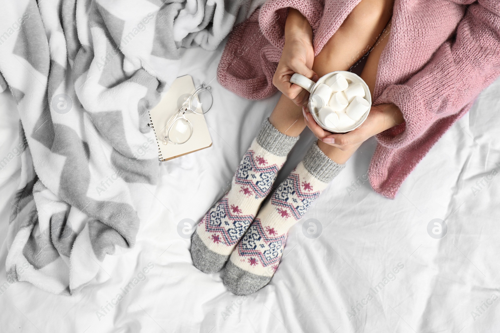 Photo of Woman with cocoa wearing knitted socks on white fabric, top view. warm clothes