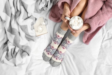 Woman with cocoa wearing knitted socks on white fabric, top view. warm clothes