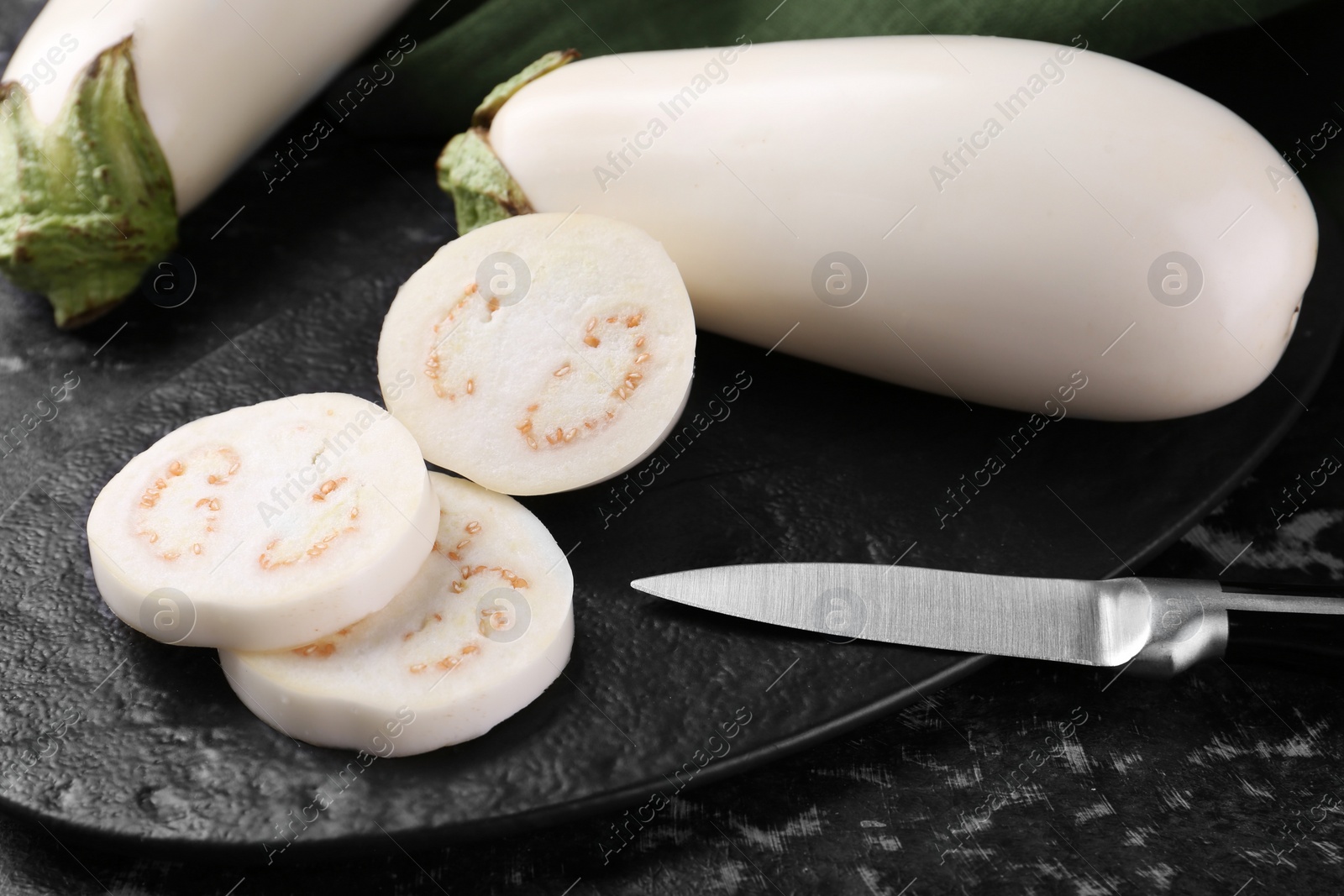 Photo of Board, raw white eggplants and knife on black textured table, closeup