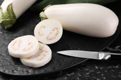 Photo of Board, raw white eggplants and knife on black textured table, closeup
